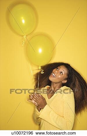 Stock Photograph - Portrait of smiling young African-American adult woman on yellow background holding balloons.. Fotosearch