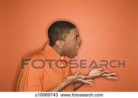 Stock Image - Side view of young African-American man on orange background expressing anger towards unseen person.. Fotosearch