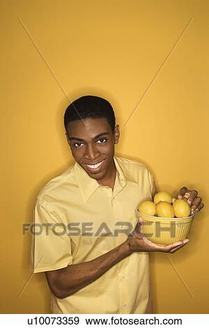 Stock Photo - Smiling young African-American man holding bowl of lemons on yellow background.. Fotosearch
