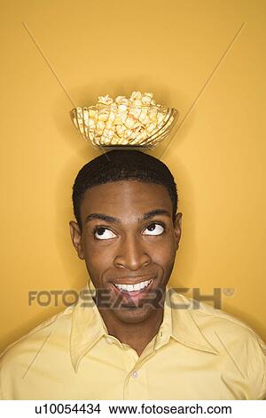 Picture - Young African-American man balancing bowl of popcorn on his head on yellow background.. Fotosearch