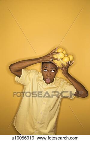 Stock Photo - Young African-American man dropping a bowl of lemons balancing on his head, on yellow background.. Fotosearch
