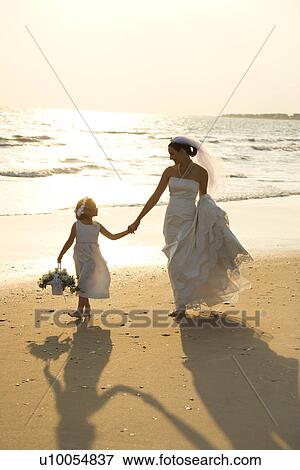 Stock Photo - Caucasian mid-adult bride and flower girl holding hands walking barefoot on beach.. Fotosearch