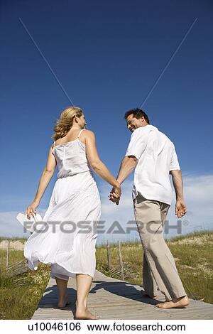 Stock Photograph - Caucasian mid-adult couple holding hands walking down beach access path.. Fotosearch