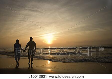 Stock Image - Caucasian mid-adult couple walking holding hands on beach at sunset.. Fotosearch