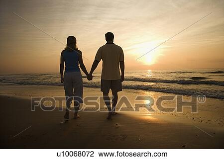 Stock Image - Caucasian mid-adult couple walking holding hands on beach at sunset.. Fotosearch