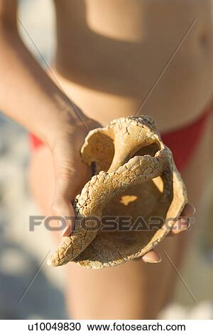 Stock Image - Caucasian mid-adult female in swimsuit holding seashell.. Fotosearch