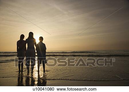 Stock Image - Caucasian mid-adult mother and teenage kids standing silhouetted on beach at sunset.. Fotosearch