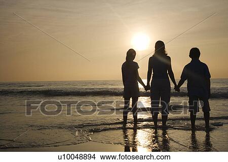Picture - Caucasian mid-adult mother and tenage kids standing silhouetted on beach at sunset.. Fotosearch