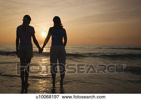 Stock Photo - Caucasian mid-adult mother teenage and daughter standing on beach at sunset holding hands.. Fotosearch