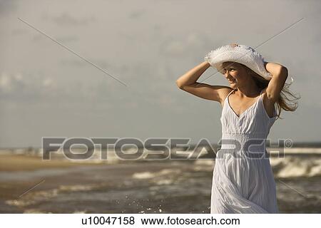 Stock Photo - Caucasian pre-teen girl standing on beach holding hat on head.. Fotosearch
