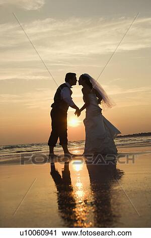Stock Image - Caucasian prime adult male groom and female bride holding hands and kissing barefoot on beach at sunset.. Fotosearch