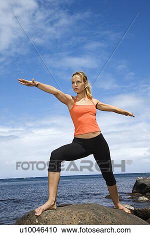 Stock Image - Caucasian young adult woman doing yoga on rocky shore.. Fotosearch