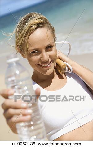 Stock Photography - Caucasian young adult woman holding jump rope and water bottle on beach.. Fotosearch