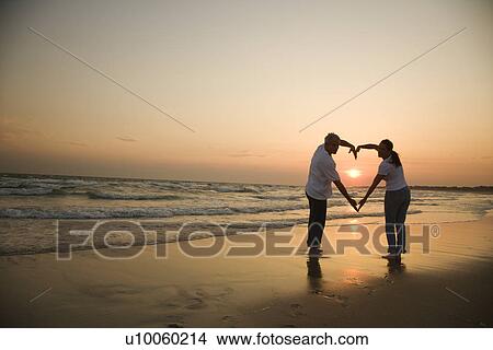 Mid-adult couple making heart shape with arms on beach at sunset. View Large Photo Image Picture - Mid-adult couple making heart shape with arms on beach at sunset.. Fotosearch