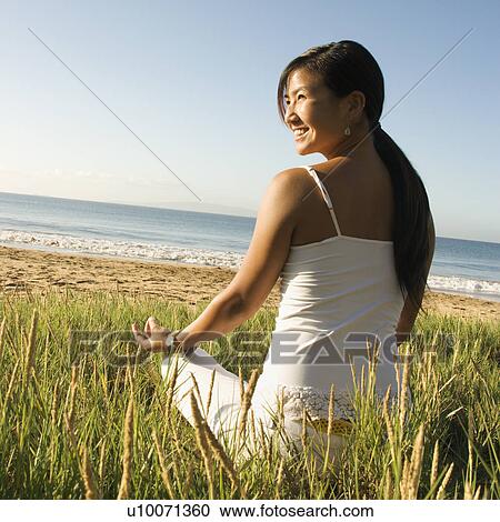Stock Image - Young Asian female sitting on beach meditating and looking off to the side.. Fotosearch