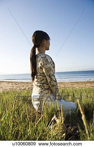 Stock Photo - Young Asian female sitting on beach looking out to ocean.. Fotosearch