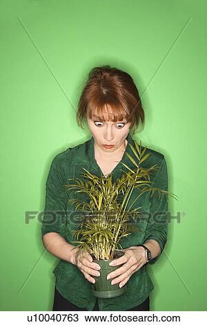 Stock Image - Caucasian mid-adult woman on green background looking at plant.. Fotosearch