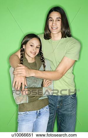 Stock Image - Portrait of Asian-American girl and teen boy standing with arms around eachother smiling against green background.. Fotosearch