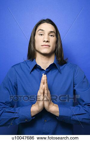Stock Photograph - Portrait of Asian-American teen boy with hands pressed together in prayer position standing against blue background.. Fotosearch