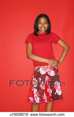 Stock Photo - Portrait of smiling African-American teen girl with hands on her hips standing in front of red background.. Fotosearch