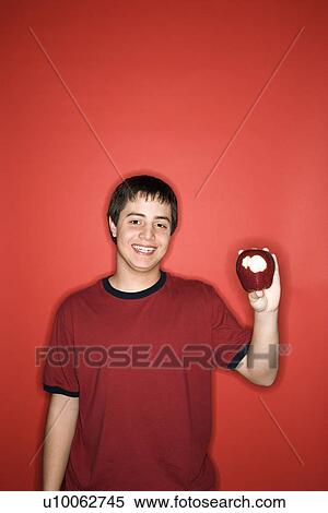 Stock Photography - Portrait of smiling Caucasian teen boy holding partially eaten apple against red background.. Fotosearch