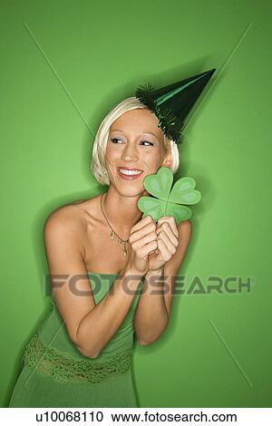 Stock Image - Portrait of smiling young adult Caucasian blond woman on green background wearing party hat and holding shamrock for Saint Patricks Day celebration.. Fotosearch