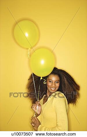 Portrait of smiling young African-American adult woman looking sweet on yellow background holding balloons. View Large Photo Image Stock Photo - Portrait of smiling young African-American adult woman looking sweet on yellow background holding balloons.. Fotosearch