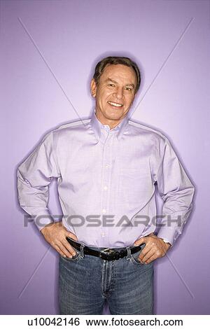 Stock Photograph - Smiling middle-aged Caucasian man with hands on hips wearing purple clothing on purple background.. Fotosearch