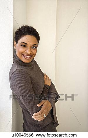 Stock Photograph - African-American woman standing leaning against wall smiling looking at viewer with arms crossed.. Fotosearch