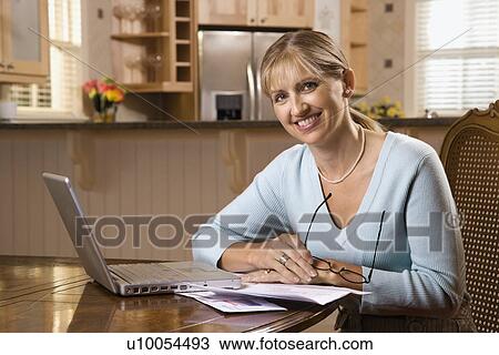 Stock Image - Caucasian mid-adult woman paying bills on laptop computer looking at viewer and smiling.. Fotosearch