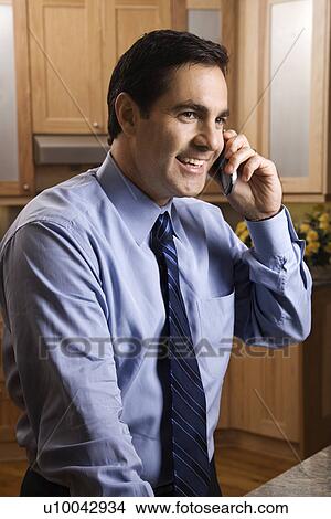 Picture - Mid-adult Caucasian male smiling and holding cell phone while standing in kitchen.. Fotosearch