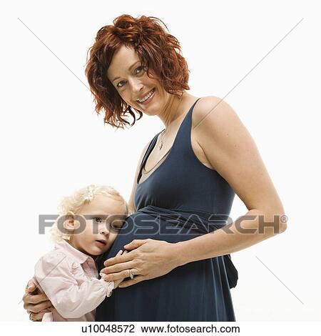 Stock Image - Caucasion mid-adult attractive pregnant smiling woman kneeling in front of female toddler who is pressing ear against belly.. Fotosearch