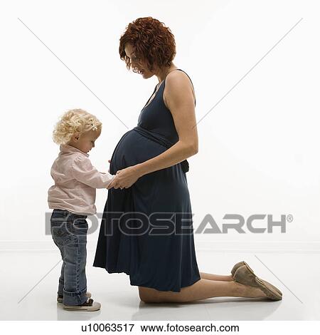 Stock Photo - Caucasion mid-adult attractive pregnant woman kneeling in front of female toddler who is touching and looking at her belly.. Fotosearch