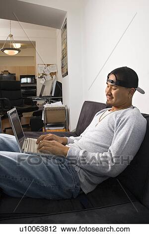 Stock Image - Asian young adult man sitting on sofa wearing cap backwards and typing on laptop.. Fotosearch