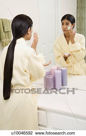 Stock Image - Asian/Indian young woman looking in mirror applying facial scrub.. Fotosearch