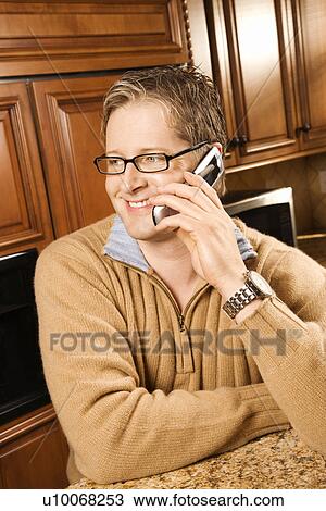 Stock Image - Caucasian man smiling and talking on cellphone while leaning on kitchen counter.. Fotosearch