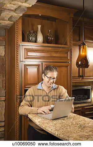 Stock Image - Caucasian man working on laptop computer in kitchen.. Fotosearch