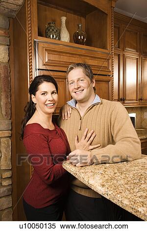 Stock Photography - Caucasian woman and Caucasian man with arms around each other leaning on kitchen counter and looking at viewer.. Fotosearch