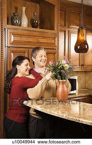 Picture - Caucasian woman leaning on Caucasian man while he arranges flowers in vase in kitchen.. Fotosearch
