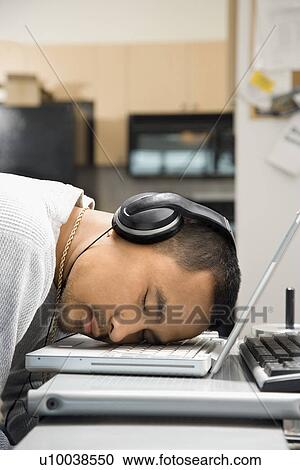 Stock Image - Close-up of Asian young adult man sleeping with head on laptop keyboard and wearing headphones.. Fotosearch