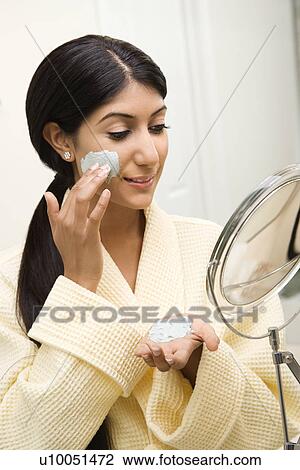 Stock Image - Smiling Asian/Indian young woman looking in mirror and applying facial scrub.. Fotosearch