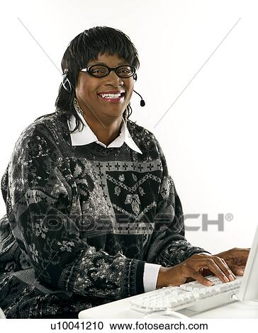Stock Image - African-American mid-adult woman wearing headset typing on computer.. Fotosearch