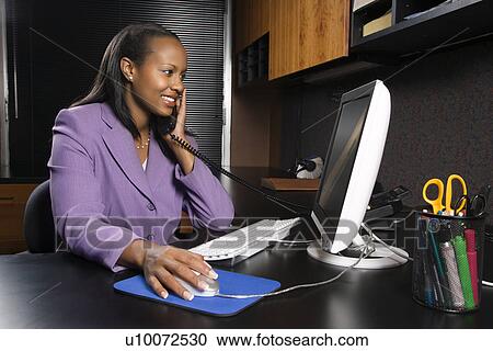 African-American young adult smiling business woman talking on phone and working at computer in office. View Large Photo Image Stock Image - African-American young adult smiling business woman talking on phone and working at computer in office.. Fotosearch
