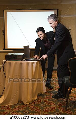 Stock Image - Asian prime adult businessman helping prime adult Caucasian businessman give presentation.. Fotosearch