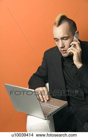 Caucasian man in suit with mohawk typing on laptop and talking on cellphone against orange background. View Large Photo Image Picture - Caucasian man in suit with mohawk typing on laptop and talking on cellphone against orange background.. Fotosearch