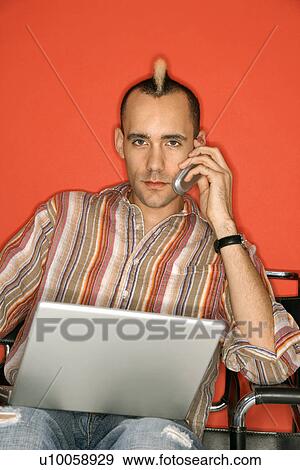 Stock Photo - Caucasian man with mohawk holding laptop talking on cellphone against red background.. Fotosearch