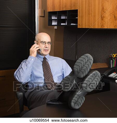 Caucasian middle-aged businessman in office sitting with feet on desk talking on cellphone. View Large Photo Image Picture - Caucasian middle-aged businessman in office sitting with feet on desk talking on cellphone.. Fotosearch
