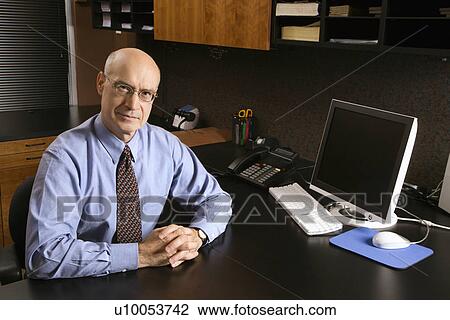 Stock Image - Caucasian middle-aged businessman sitting at desk in office.. Fotosearch