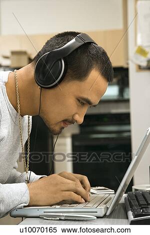 Close-up side view of Asian young adult man leaning over laptop typing on keyboard wearing headphones. View Large Photo Image Stock Photography - Close-up side view of Asian young adult man leaning over laptop typing on keyboard wearing headphones.. Fotosearch