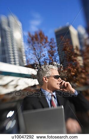 Prime adult Caucasian man in suit sitting at patio table outside with laptop talking on cellphone wearing sunglasses with buildings in background. View Large Photo Image Stock Image - Prime adult Caucasian man in suit sitting at patio table outside with laptop talking on cellphone wearing sunglasses with buildings in background.. Fotosearch